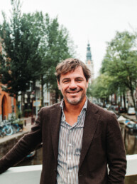 Maarten de Gruyter poseert in een bruin jasje op een brug in Amsterdam