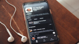 Foto van een smartphone op een tafel met iPhone oortjes links ervan. Op het scherm is de app van Spotify te zien met daarop 'The Joe Rogan Experience'