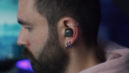 Close-up van een oor van een man met een baard. De man heeft oordopjes in zijn oor en luistert naar een podcast. De man heeft ook drie oorringen in zijn oor.